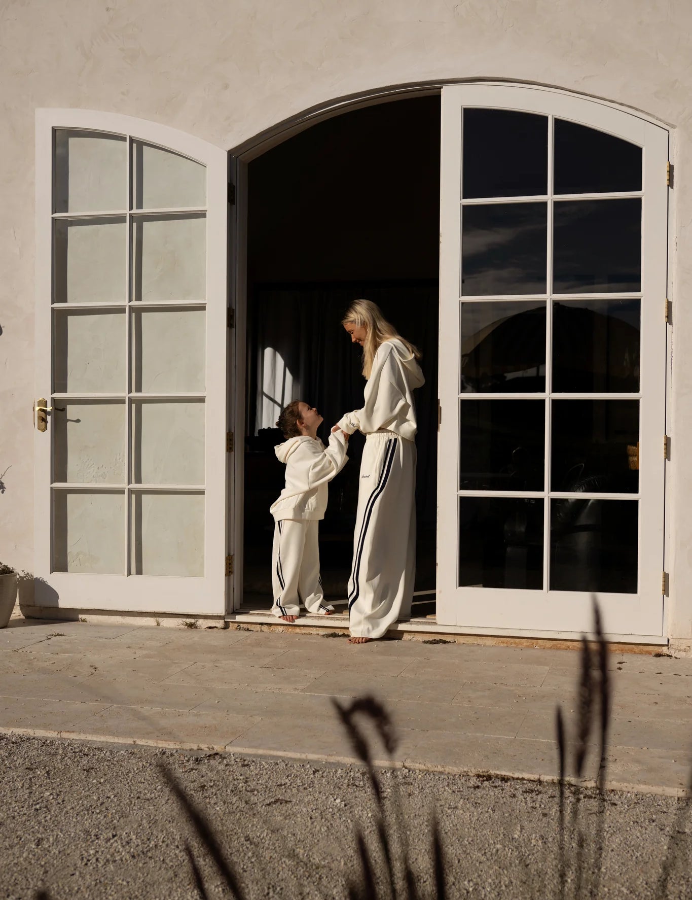 Mother and child wearing matching cream loungewear standing in a sunlit doorway – stylish family fashion by Wild West Sisters