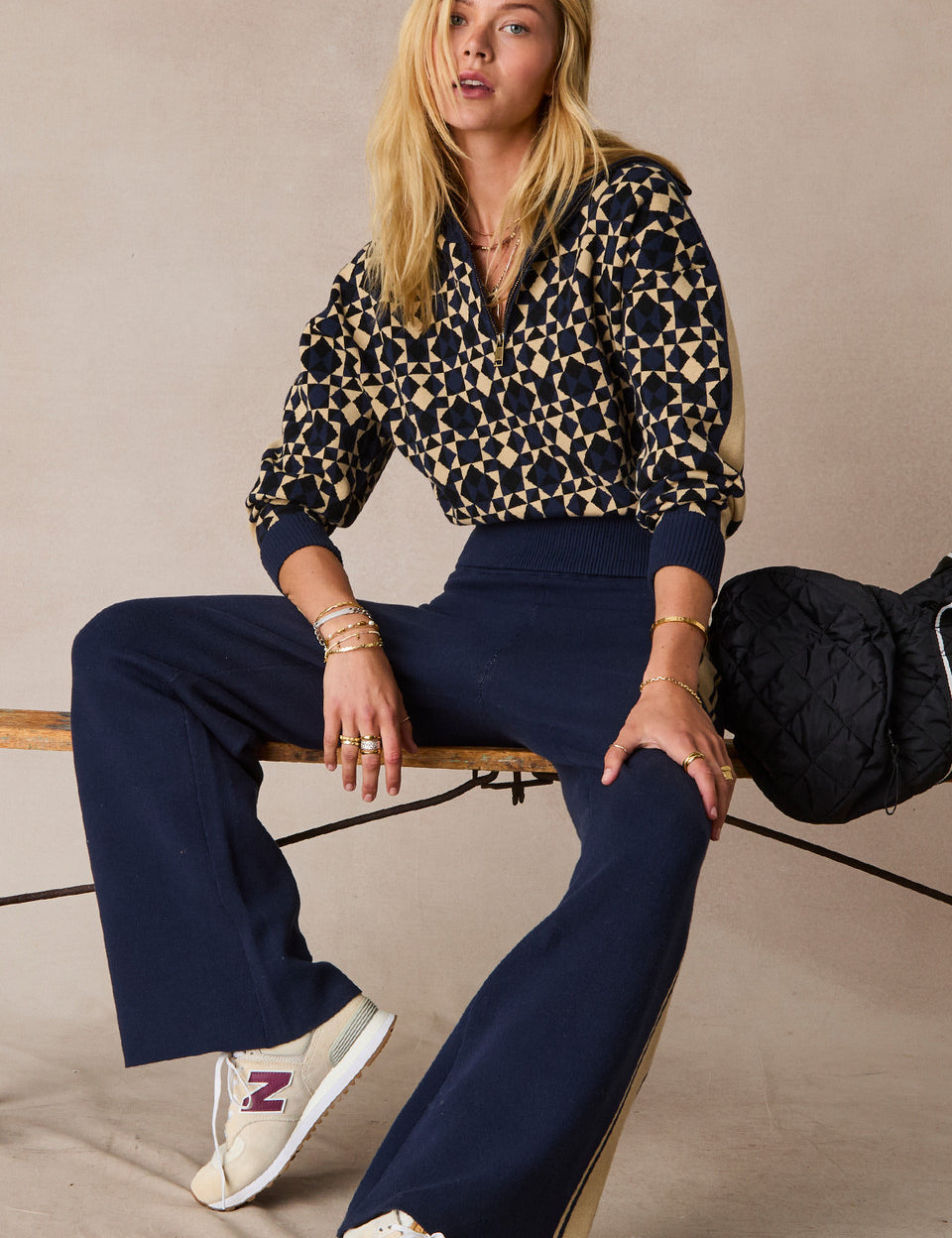 Woman sitting on a wooden stool wearing a patterned top and navy pants with a beige background