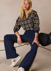 Woman sitting on a wooden stool wearing a patterned top and navy pants with a beige background