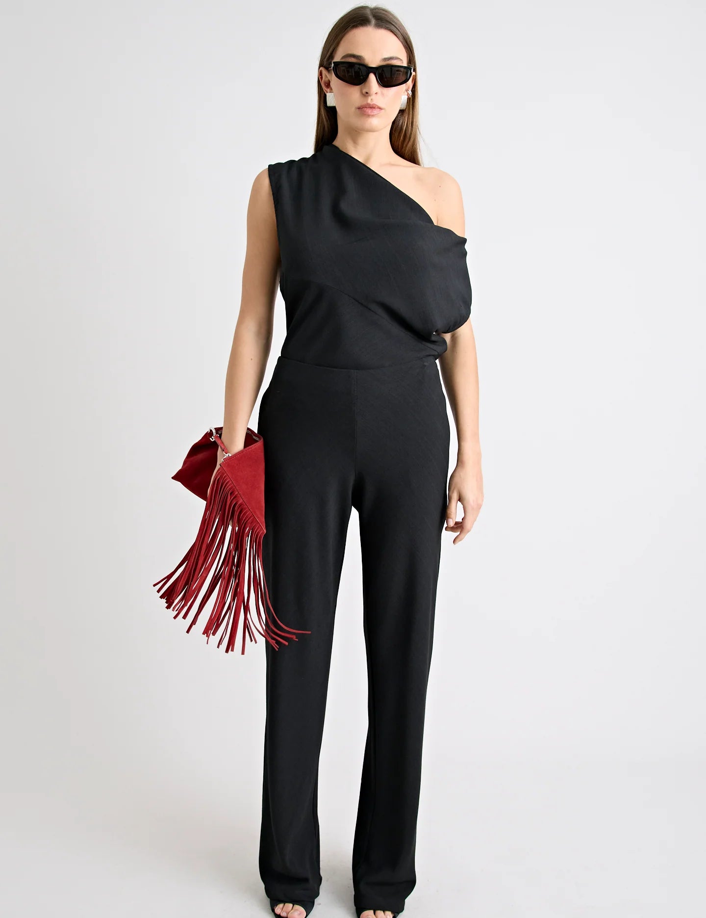 Woman wearing a black one-shoulder top and pants with a red fringed bag on a white background