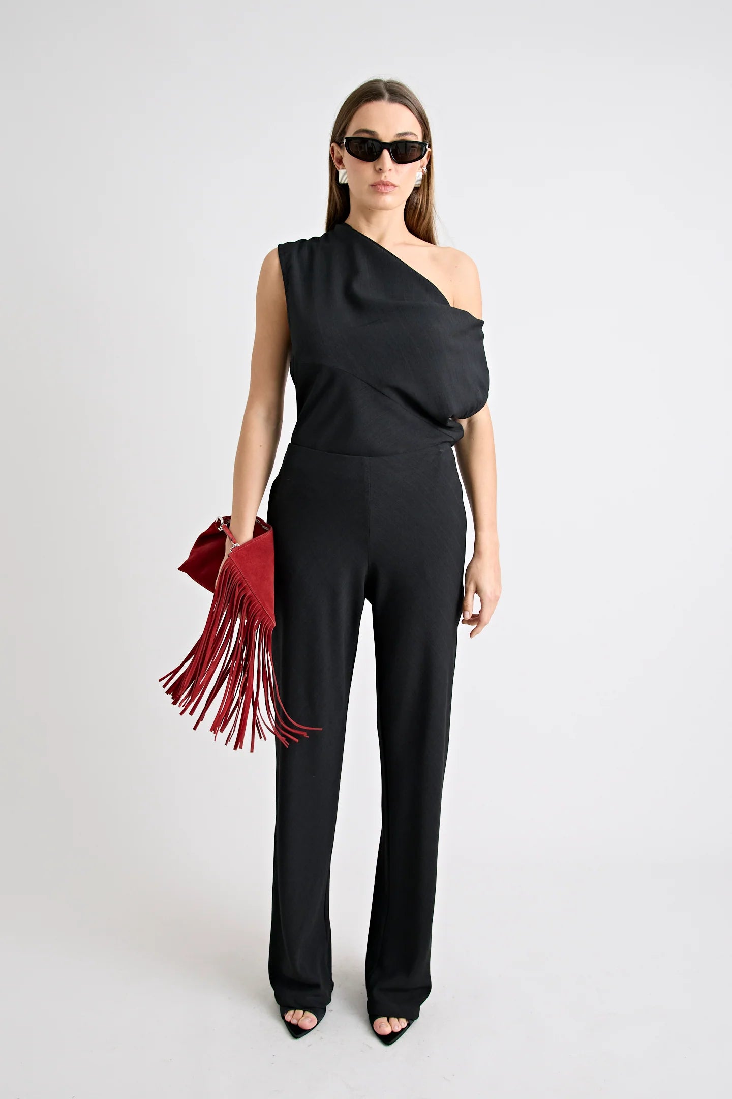 Woman wearing a black one-shoulder top and pants with a red fringed bag on a white background